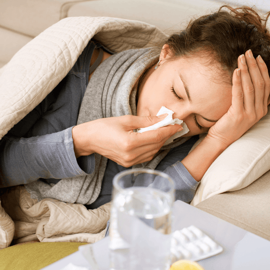 Woman feeling unwell with a tissue, resting under a blanket beside a glass of water.